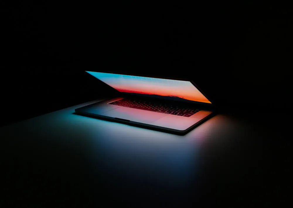 An image of a laptop in the dark, half closed, with the backlight from the screen illuminating its keyboard and the surrounding surface.