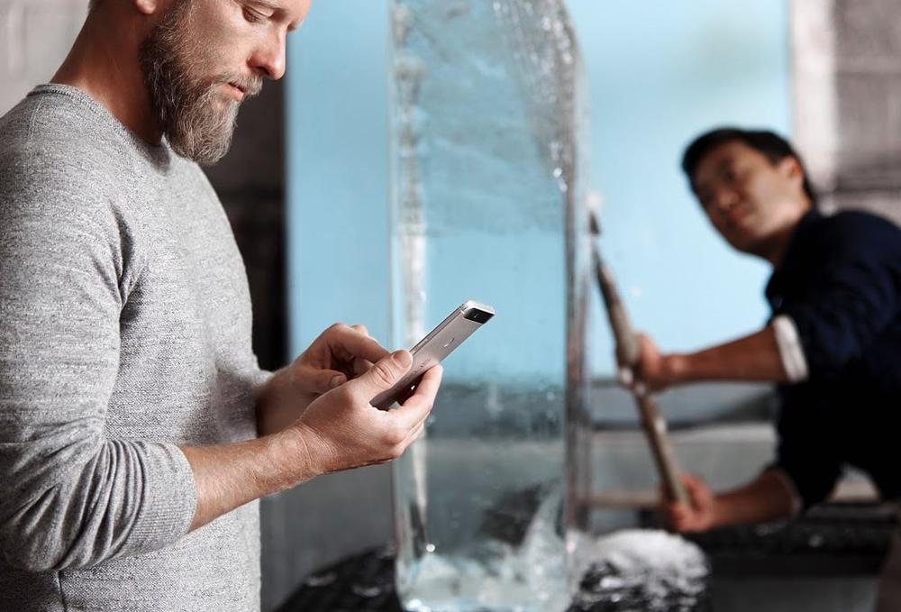An image of a man using a Nexus smartphone, with another man creating an ice sculpture behind him for some reason, out of focus.