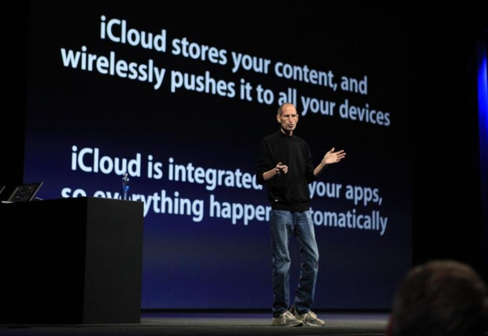 Photograph of Steve Jobs on stage delivering a keynote, flanked by a slide with text reading “iCloud stores your content, and wirelessly pushes it to all of your devices. iCloud is integrated with your apps, so everything happens automatically.”
