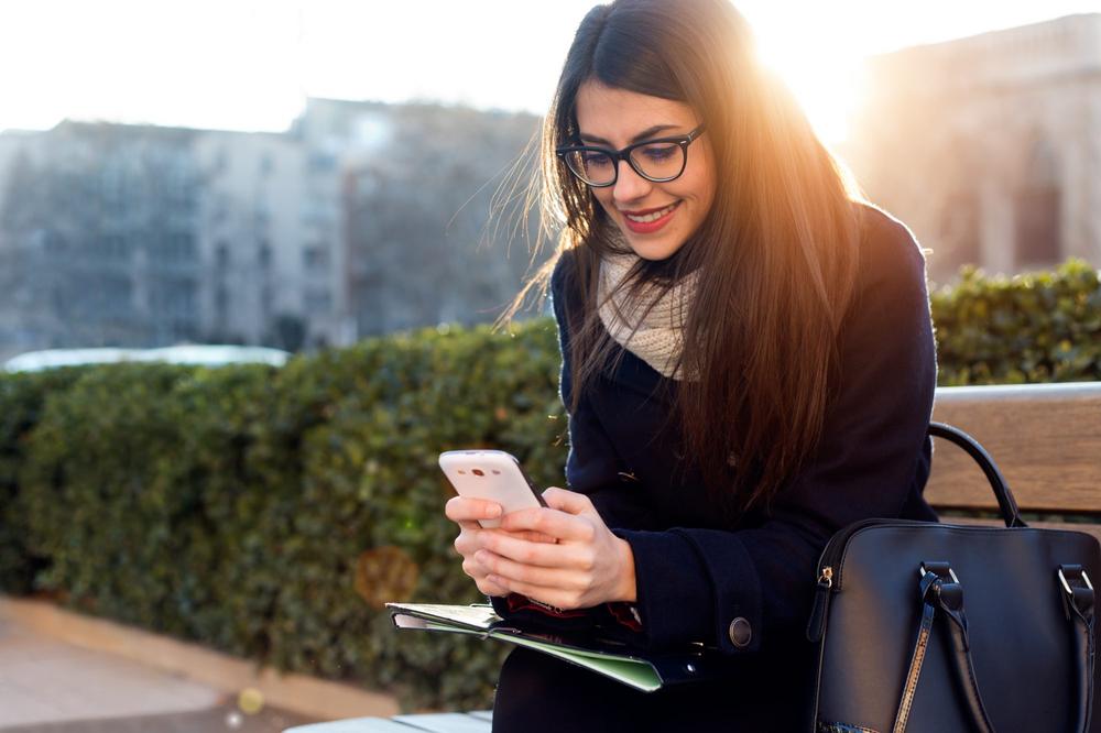 Photograph of a woman using an embarrassingly ancient Android handset, sitting outdoors, smiling.