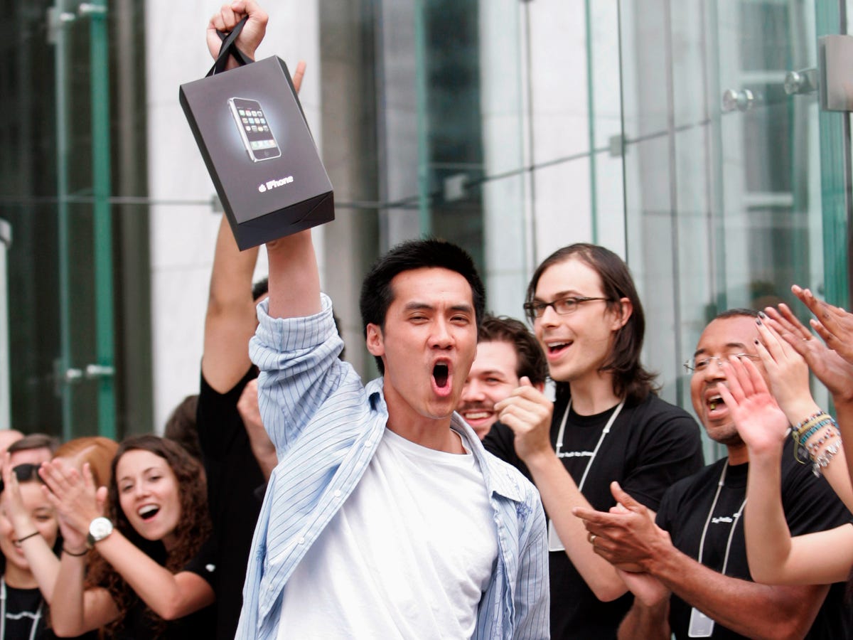 Photograph of presumably the first iPhone customer, a man triumphantly holding up his first-generation iPhone bag outside the Fifth Avenue Apple Store, with Apple retail employees looking on and clapping, in New York City in June 2007.